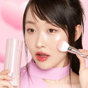 Close-up of a young woman applying blush with a soft brush, featuring dewy K-beauty skin, glossy lips, and a fresh pink-toned makeup look against a pastel background.