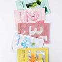 Flat lay of colorful K-beauty sheet mask packets arranged in a fan, featuring pastel packaging with bold numbers and ingredients like aloe, vitamin C, and soothing botanicals on a clean white background.