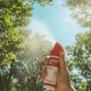 Hand holding a Purito hypochlorous acid facial mist spray outdoors, releasing a fine mist against a bright blue sky and sunlit green trees, highlighting a fresh, refreshing K-beauty skincare moment.