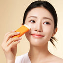 Smiling woman with radiant, dewy K-beauty skin applying a balm stick to her cheek, showcasing a natural glow and minimal makeup against a soft beige background.