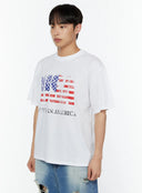 Men's US Flag Graphic Tee IU516 in white, three-quarter side angle on model, showing oversized drop-shoulder fit and distressed USA flag graphic with Made in America lettering, worn with ripped blue jeans.