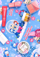 A flat-lay of a colorful cosmetic advent calendar or gift box featuring cartoon bunnies, berries, and numbered windows on a bright blue background. Visible products include pink and red makeup items, a jar with yellow contents, and a round compact labeled 'ALL CLEAN BLACK' by 'missmeow'.